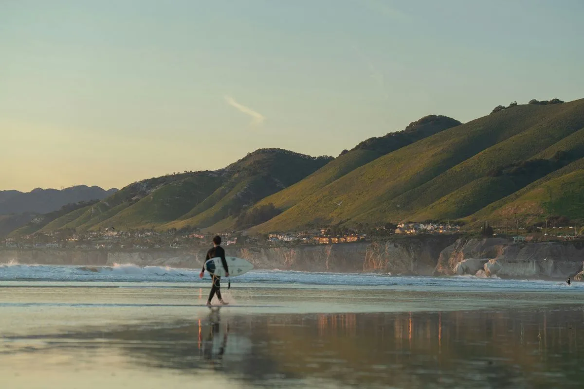 Person walking on a beach with a surfboard, representing beach college lifestyle